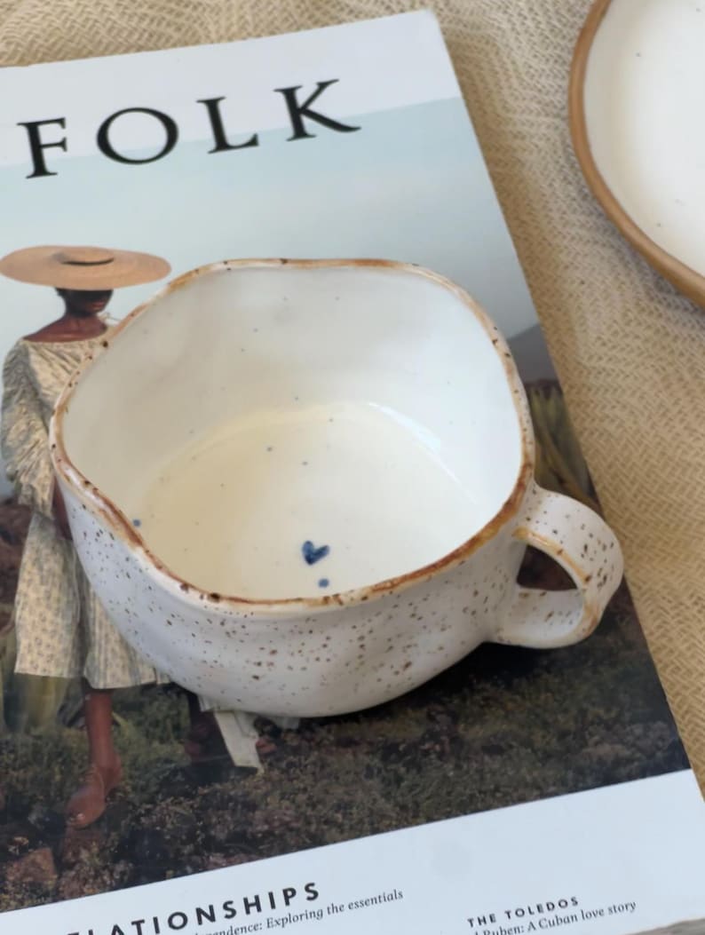 May include: A speckled white ceramic mug with a brown rim and handle. The mug has a unique, irregular shape and a small blue heart detail. The mug is placed on a magazine with the word "FOLK" visible.