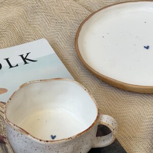 May include: A white ceramic mug and plate set with a speckled finish and a brown rim. The mug and plate each feature a small blue heart design. The word "LK" is visible on a magazine in the background.