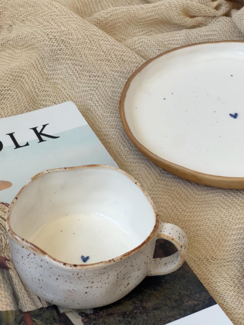 May include: A white ceramic mug and plate set with a speckled finish and a brown rim. The mug and plate each feature a small blue heart design. The word "LK" is visible on a magazine in the background.