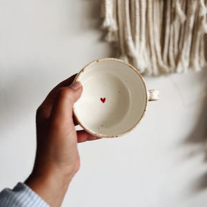 Könnte beinhalten: Eine weiße Keramiktasse mit einem kleinen roten Herz, das innen bemalt ist. Die Tasse hat eine gesprenkelte Glasur und einen Henkel.