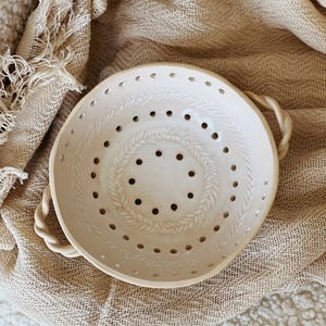 May include: A white ceramic colander with a woven design and holes for draining. The colander has two rope handles.