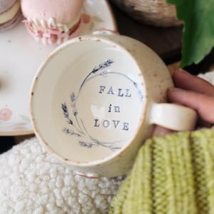 May include: Cream ceramic mug with a speckled finish. The interior has the words "FALL in LOVE" in dark blue, encircled by a floral wreath. The mug has a handle and is set against a soft, textured background.