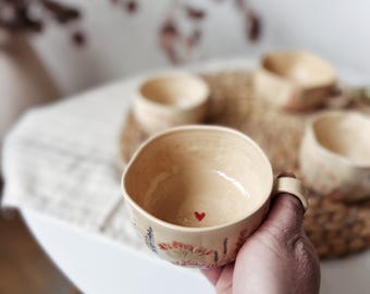 May include: A beige ceramic teacup with a small red heart painted inside. The cup has a handle and is decorated with floral and lavender designs. Other similar cups are visible in the background, suggesting a set.