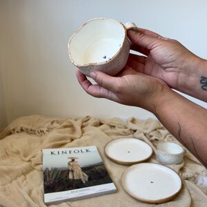 May include: A handmade ceramic mug with a unique, irregular shape, speckled exterior, and a small handle. The interior is white, and the rim has a brown accent. Two small plates and a small bowl are also visible, along with a Kinfolk magazine.