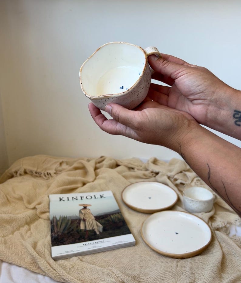 May include: A handmade ceramic mug with a unique, irregular shape, speckled exterior, and a small handle. The interior is white, and the rim has a brown accent. Two small plates and a small bowl are also visible, along with a Kinfolk magazine.