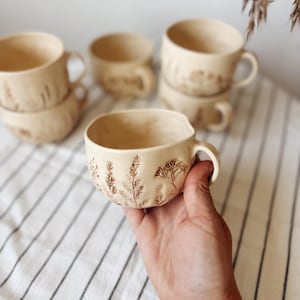 Könnte beinhalten: Ein Set aus drei Keramikbechern mit gesprenkelter, matter Oberfläche. Jeder Becher hat eine andere Farbe: hellblau, cremefarben und grau. Die Becher haben ein schlichtes, modernes Design und einen bequemen Griff.