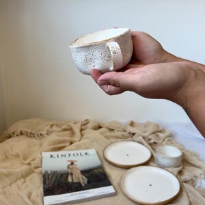 May include: A speckled white ceramic mug with a handle is held in a person's hands. The mug is part of a set that includes two small plates and a small bowl. A copy of "Kinfolk" magazine is also in the image.