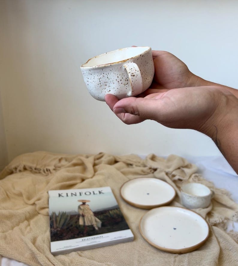May include: A speckled white ceramic mug with a handle is held in a person's hands. The mug is part of a set that includes two small plates and a small bowl. A copy of "Kinfolk" magazine is also in the image.