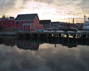 May include: A scenic waterfront view featuring red buildings, a wooden pier, and calm water reflecting the sky. A black truck and other vehicles are parked on the pier. The sky is a mix of orange and gray.