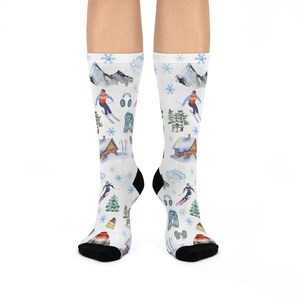 May include: White socks with a winter ski scene pattern. The socks feature illustrations of skiers, snow-covered cabins, mountains, snowflakes, and trees. The heels and toes are black.