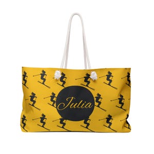 May include: Yellow tote bag with a repeating black skier pattern. The bag has a large black circle with the name "Julia" in gold script. It features white rope handles.