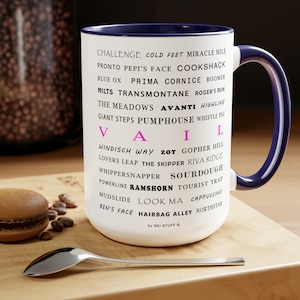May include: White ceramic coffee mug with a blue rim and handle. The mug features a black and pink design with the word "Vail" in large pink letters and a list of ski runs and locations in smaller black letters. The text "by Ski Stuff" is printed in black at the bottom of the mug.