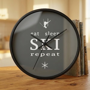 May include: A round wall clock with a black frame and a gray face. The clock face features the words "eat sleep SKI repeat" in white, along with a skier graphic and a snowflake. The clock is on a wooden surface.