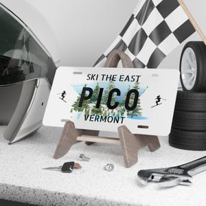 May include: A white license plate with the text "SKI THE EAST" and "PICO VERMONT" over a mountain scene. The plate is displayed on a wooden easel, with a helmet, tires, and a checkered flag in the background.
