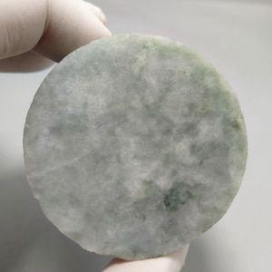 Puede incluir: Una piedra de jade redonda, de color verde pálido, con un aspecto moteado. La piedra tiene una superficie lisa y pulida y una textura natural. El jade está sostenido por una mano enguantada, mostrando su tamaño y forma. El color varía de verde claro a oscuro.