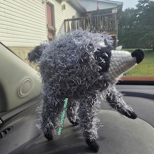May include: A handmade crochet raccoon plush toy featuring a fuzzy grey body, a white pointed snout, black eye patches, and black paws. A small green tag with the text shiny_lunchbox_studio is attached to the side of the raccoon's body.