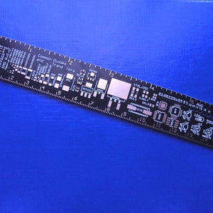Könnte beinhalten: Ein schwarzes Lineal mit weißen Markierungen und einem Leiterplatten-Design. Das Lineal ist 38 cm lang und hat Markierungen für Zoll und Zentimeter. Das Leiterplatten-Design umfasst gängige Dioden und Transistoren, einen Quarzoszillator und einen 5 AWG-Draht.