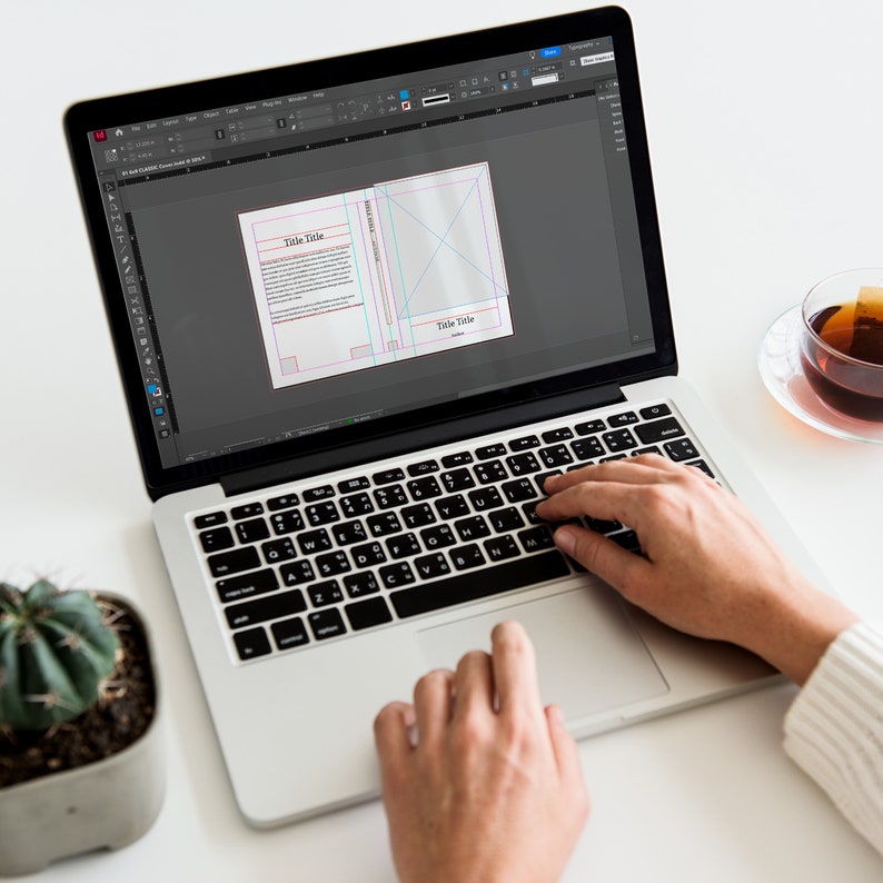 May include: A person is using a laptop computer to design a book cover. The cover has the text "Title Title" on it. The laptop is on a white table with a cactus plant and a cup of tea.