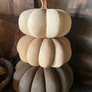 May include: A stacked pumpkin decoration with three sections. The top two sections are light beige, and the bottom section is a darker brown. The pumpkin has a textured appearance and a curved, brown stem. The decoration is likely for fall or autumn.