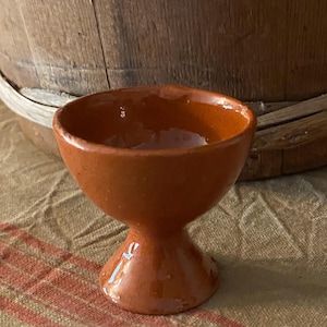 May include: A small, glazed terracotta egg cup with a rounded bowl and a pedestal base. The egg cup is a warm, reddish-brown color and sits on a striped fabric. A wooden barrel is in the background.