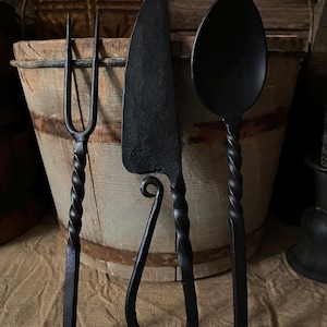 May include: Three black metal utensils with twisted handles. A fork, knife, and spoon are displayed against a wooden background.