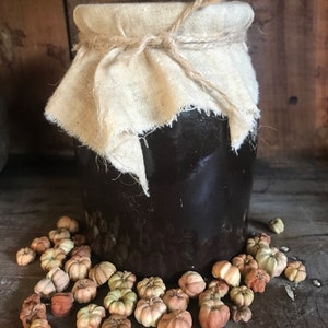 May include: A brown ceramic jar with a cloth lid tied with twine, surrounded by small, light brown, dried flower buds.