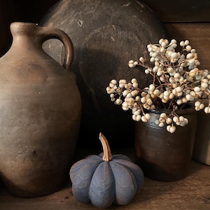 May include: A rustic still life featuring a large brown jug with a handle, a small dark blue fabric pumpkin, and a small brown pot filled with white berry branches. The items are arranged on a wooden surface.