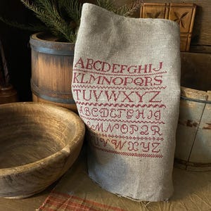 Könnte beinhalten: Ein Leinen-Sampler mit dem Alphabet, das in roter Farbe gestickt ist. Der Sampler ist gefaltet und liegt auf einer Holzoberfläche mit einer Holzschüssel und einer Holzkiste.