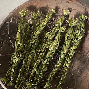 May include: Braided green and yellow dried herbs, tied with black bands, rest in a rustic wooden bowl. The herbs have a textured appearance and are arranged in a decorative pattern. The bowl has a natural, aged look.