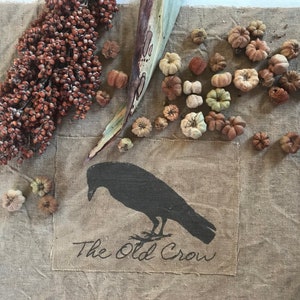 May include: A burlap fabric with a black crow silhouette and the text "The Old Crow". The fabric is decorated with dried gourds and a dried stalk of sorghum.