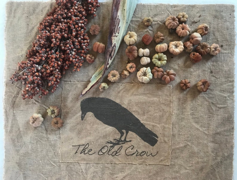 May include: A burlap fabric with a black crow silhouette and the text "The Old Crow". The fabric is decorated with dried gourds and a dried stalk of sorghum.