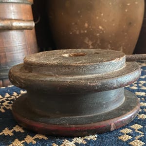 以下が含まれることがあります： 中央に穴と金属製のハンドルが付いた、風化した濃い茶色の木製スプール。 スプールは、エッジにわずかに赤みを帯びた多層設計になっています。 背景には木製の樽やその他のアンティーク品が含まれています。