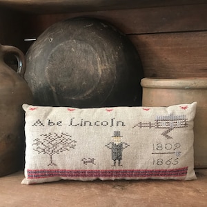 May include: A vintage-style decorative pillow with cross-stitch embroidery featuring the name "Abe Lincoln", a tree, a dog, and the years 1809-1865. The pillow has a red and black border and is displayed with antique pottery.