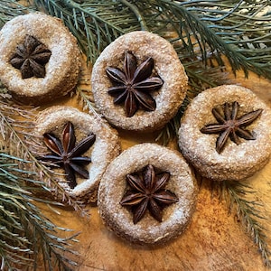 May include: Five round, light brown cookies with a dusting of white powder and a star anise center. The cookies are arranged on a wooden surface with green sprigs. A close-up shot of the baked goods.