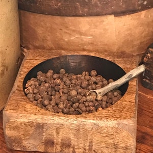 May include: A wooden bowl filled with black peppercorns and a small wooden spoon. The bowl is square with rounded edges and has a dark brown stain.