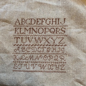 May include: A beige linen fabric with the alphabet embroidered in brown thread. The letters are in a simple, blocky font and are repeated twice.