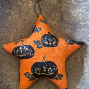 May include: A star-shaped fabric ornament in bright orange featuring a repeating print of dark navy blue jack-o'-lanterns and black leaves. The ornament includes a thin black string loop attached to the top point for hanging purposes.