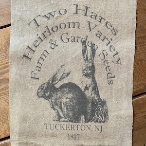 May include: A beige linen fabric with black text and a black and white illustration of two rabbits. The text reads "Two Hares Heirloom Variety Farm & Garden Seeds Tuckerton, NJ 1817".