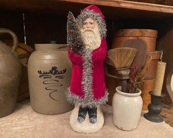 Op de afbeelding: Een vintage Santa Claus beeldje in een rode fluwelen jas met zilveren afwerking, met een kleine kerstboom. Het beeldje heeft een witte baard en staat op een besneeuwde basis. Andere items zijn een steengoed pot en een kaars.