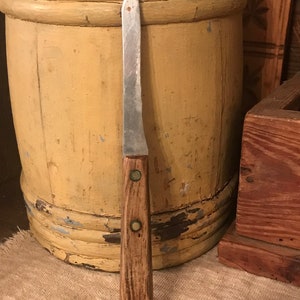 May include: A vintage kitchen knife with a wooden handle and a silver blade. The knife is resting against a yellow painted wooden barrel.