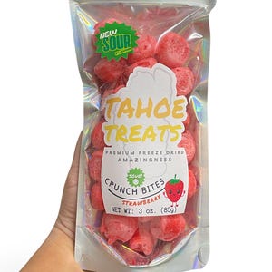 Può includere: Un sacchetto trasparente olografico pieno di crunch bites di fragole liofilizzate rosse. La confezione presenta un'etichetta con la scritta "TAHOE TREATS" e "SOUR CRUNCH BITES STRAWBERRY". Il peso netto è di 85g.