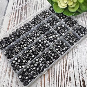 May include: A clear plastic organizer filled with black round beads, each with a white letter or number. The beads are arranged in individual compartments. A green succulent is visible in the background. Ideal for jewelry making and crafting.