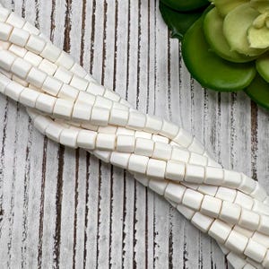 May include: A strand of white square beads made of bone. The beads are smooth and have a slightly irregular shape. The beads are strung on a white cord.
