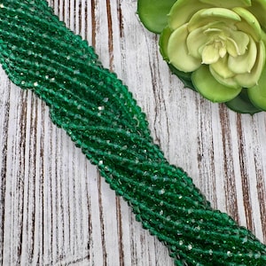May include: Strands of faceted, emerald green beads. The beads are strung together, creating a flowing, textured appearance. The beads are displayed on a white and brown wooden surface, with a green succulent in the background.
