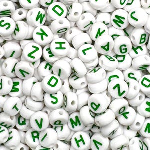 May include: A close-up of a pile of white plastic letter beads with green lettering. The beads are round and have a hole in the center for stringing.