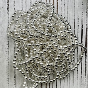 May include: A tangled strand of shiny, silver-colored beads. The beads are small, round, and evenly spaced along a thin string. The beads are reflective, creating a bright, metallic appearance. The background is a white, weathered wooden surface.