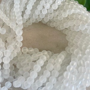 May include: Close-up of a strand of white, frosted glass beads. The beads are round and uniform in size, strung together to form a continuous line. The beads have a matte finish, creating a soft, diffused appearance. Ideal for jewelry making.