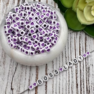 May include: White square beads with purple heart designs are piled in a marble bowl. The beads are next to a string of white square beads that spell out "BOSS LADY" with purple heart accents. The background is a white wooden surface.
