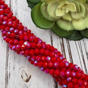 May include: Close-up of strands of bright red, faceted beads with iridescent flashes. The beads are round and tightly packed. A green succulent and a white-painted wooden surface are in the background.