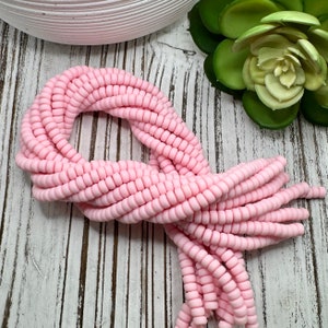 May include: A collection of light pink, spiral-shaped beads, arranged in a looped pattern. The beads are uniform in size and shape, with a matte finish. The background features a white, weathered wooden surface and a green succulent.